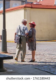 Kaunas, Lithuania - 05 19 2017: Stylish Couple Of Elderly People Are Happy Together. A Woman With Red Hair And Hat And A Grizzly Man With Sunglasses Are Standing On The Stone Pavement And Chatting.