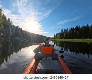 Kayaking Through Forested River in Summer Afternoon. The sun shines brightly in the blue sky, casting reflections on the water. The kayak is orange and the person is wearing an orange life vest.