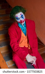 KAZAKHSTAN, KOSTANAY - OCTOBER 21, 2019: A Man With A Joker Makeup And In A Red Suit Sits On The Stairs And Smokes A Cigarette.