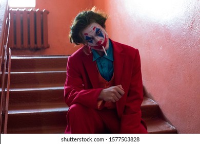 KAZAKHSTAN, KOSTANAY - OCTOBER 21, 2019: A Man With A Joker Makeup And In A Red Suit Sits On The Stairs And Smokes A Cigarette.