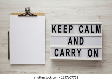 'Keep calm and carry on' words on a modern board, clipboard with blank sheet of paper on a white wooden background, top view. Overhead, from above, flat lay. 