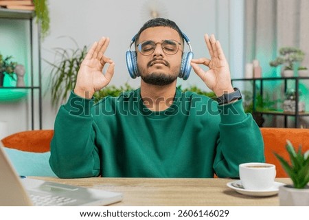 Keep calm down, relax. Indian man listening music breathes deeply, eyes closed meditating with concentrated thoughts, peaceful mind. Young Hindu Arabian guy sits at home office room table workplace
