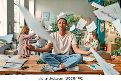 Keep calm and no stress! Young African-American man is sitting in lotus position while his colleagues are arguing nearby. Multiracial people working together in modern office. - Shutterstock ID 1958384992