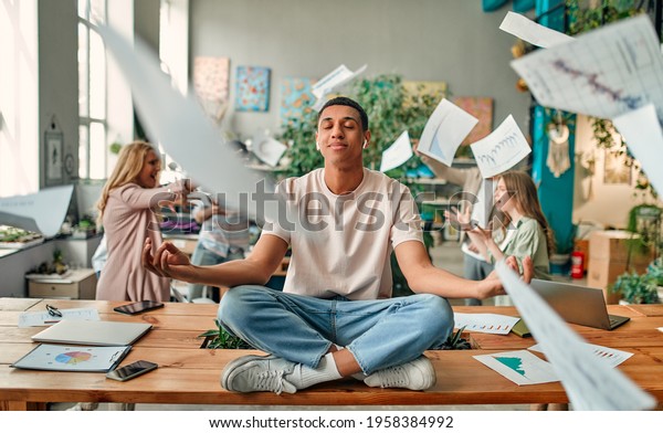 Keep calm and\
no stress! Young African-American man is sitting in lotus position\
while his colleagues are arguing nearby. Multiracial people working\
together in modern office.