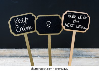 Keep Calm And Take It To The Next Level Message Written With Chalk On Mini Blackboard Labels, Defocused Chalkboard And Wooden Table In Background. Fun And Humor Concept