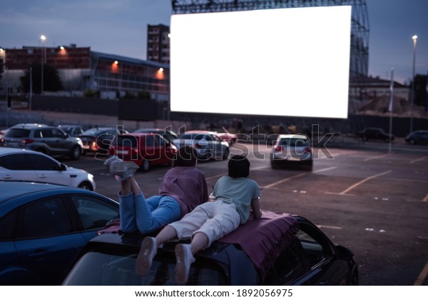 Keep calm and watch the movie. Rear view of\
two friends lying on the roof of a car, having fun while watching a\
movie in an open air cinema. Safe entertainment during coronavirus\
quarantine