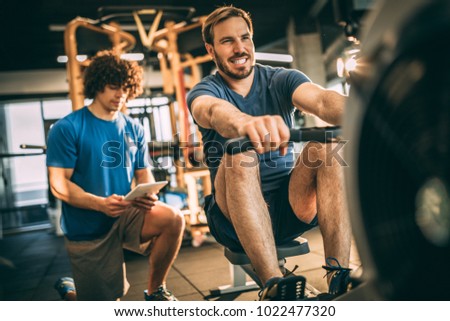 Keep going! You can do it! Mid adult man with his personal instructor in fitness center. Rowing machine.