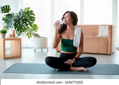 Keep hydrated. Young Hispanic woman drinking water on yoga mat at home
