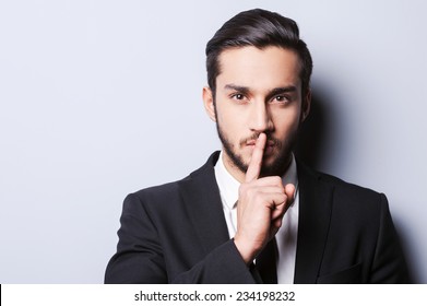 Keep my secret!  Serious young man in formalwear holding finger on lips and looking at camera while standing against grey background