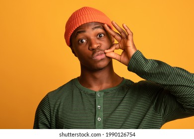 Keep Quiet. Young Black Hipster Guy In Orange Hat Making Zipping Mouth Gesture, Holding Fingers Near Lips, Funny African American Man Promising To Hold Secret, Standing On Yellow Background, Closeup