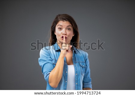 Keep a secret between us. A young female in a casual outfit stands next to a gray wall and gives a sign to be silent. Discreet relationship, hiding something from other people, speechless