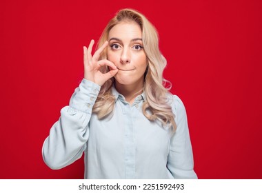 Keep the secret - keep quiet. Young woman keeps closed mouth with her fingers, isolated on red background. Studio shot - Shutterstock ID 2251592493