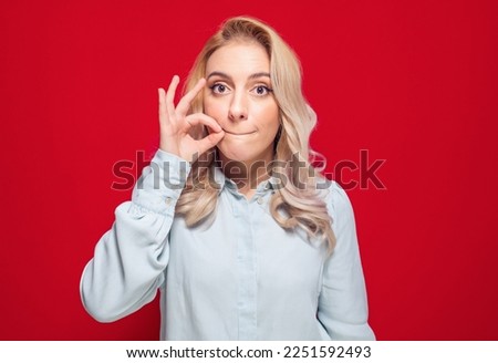 Keep the secret - keep quiet. Young woman keeps closed mouth with her fingers, isolated on red background. Studio shot