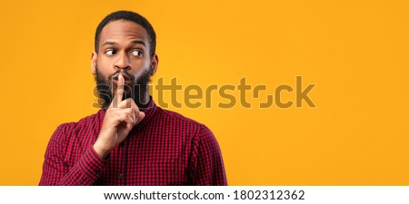 Keep Silence. Black guy making hush sign, holding finger on lips, looking aside at copy space, yellow wall, panorama