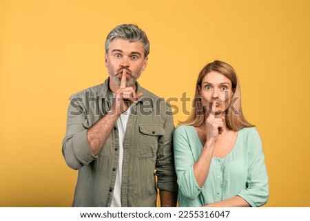 Keep silence concept. Middle aged spouses gesturing hush sign and looking at camera, standing over yellow studio background. Couple holding finger on lips, silent gesture, secret