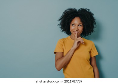 Keep silence gesture. Cheerful afro girl showing shhh sign with finger near lips, standing over pastel blue background with copy space and mysteriously looking away. Positive women emotions concept - Powered by Shutterstock