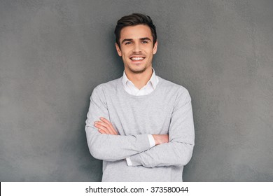 Keep smiling. Confident young man keeping arms crossed and looking at camera with smile while standing against grey background