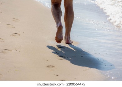 Keep Walking Concept, Selective Focus Of Bare Men's Feet With Approaching Seawave On The Shore, Low Angle Of Of A Man Walking On The Sand Beach With Footprints, Activities And Recreation In Summer.