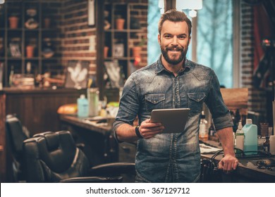 Keeping business on top with digital technologies. Cheerful young bearded man looking at camera and holding digital tablet while standing at barbershop