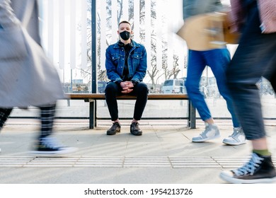 Keeping Safe Social Distance Concept. Young Hipster Man With Black Protective Face Mask Sits On Bus Stop Or Tram Station Outdoor On The Crowded Urban Street. Prevent For Coronavirus Outbreak.