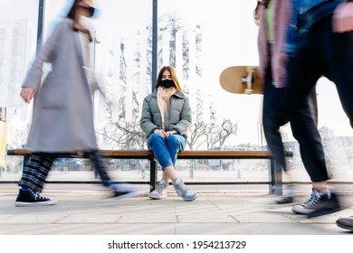 Keeping safe social distance concept. The woman with medical face mask waiting for transport sitting on bus stop on the crowded street outdoor. Portrait with motion blurred foreground.