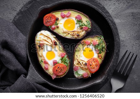 Keto diet dish: Avocado boats with ham cubes, quail eggs, cheese and cress sprouts on cast iron skillet with towel on dark background, top view