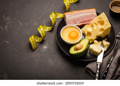 Keto foods as butter, olive oil, fried egg, avocado, fat meat bacon, cheese for ketogenic diet on black ceramic plate, measure tape on dark rustic stone background, products for weight loss copy space