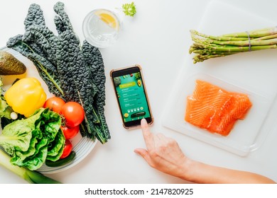 Keto meal diet planning. Woman holding phone with keto menu in mobile app on table with fresh ingredient, fresh vegetables and salmon fish. Ready to cook. Healthy lifestyle, weight loss. Recipe box.