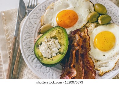 Ketogenic breakfast with bacon and eggs.Delicious breakfast with bacon and eggs From above plate with appetizing fried eggs olives Brazil nuts and stuffed avocado on white table. Ketogenic diet
