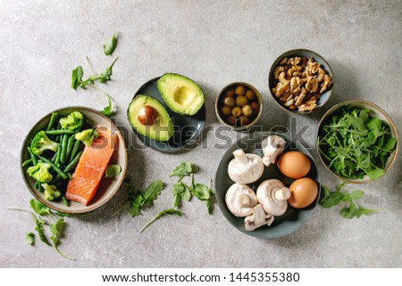 Ketogenic diet ingredients for cooking dinner. Raw salmon, avocado, broccoli, bean, olives, nuts mushrooms, eggs in ceramic bowls. Grey texture background. Flat lay, space