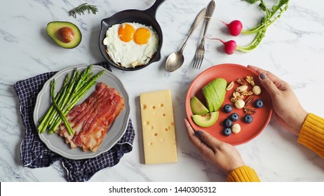 Ketogenic diet meal preparation female hands serving red plate with avocado, blueberry, Brazil nut, macadamia, pecan, walnut on marble table with fried eggs, bacon, roasted asparagus and cheese. - Shutterstock ID 1440305312
