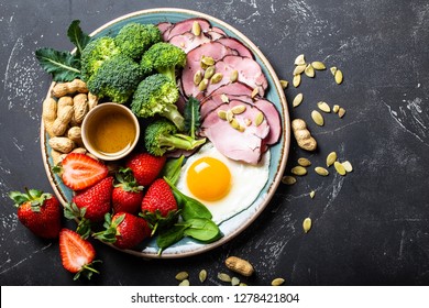 Ketogenic low carbs diet concept, top view. Plate on stone black background with keto foods: egg, meat, olive oil, broccoli, berries, nuts, seeds, spinach. Healthy fats, clean eating for weight loss