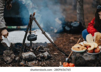 A kettle hangs over a small campfire as people prepare bread and fruit at a forest campsite. The scene shows outdoor cooking and casual camping food prep with smoke rising from the fire.
