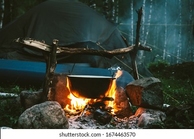 Kettle On Fire Near Tent In Forest At Night. Beautiful Campfire In Tourist Camp Into Wild. Survival In Taiga. Cauldron Above Bonfire. Smoke From Fire Among Trees. Cooking Over Campfire.