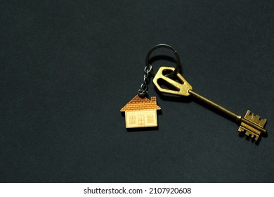 Key Chain In The Shape Of House With Key On A Black Background. Building, Design, Project, Moving To New Home, Mortgage, Deposit, Rent And Purchase Real Estate. Copy Space