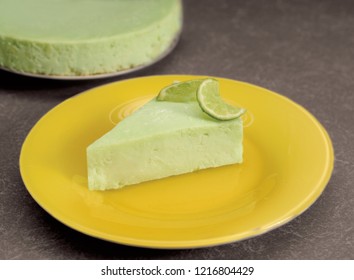 Key Lime Pie Cheesecake Slice With Lime Slice On Top, On Yellow Plate With Whole Pie In Background.