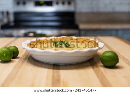 Key lime pie in a white pie dish on a wooden kitchen counter with a twist. Key limes next to pie dish. 