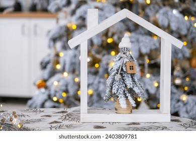 Key on Christmas tree and tiny house on cozy home with Christmas decor on table of festive white kitchen. Gift for New Year. Insurance, moving to new house, mortgage, rent and real estate