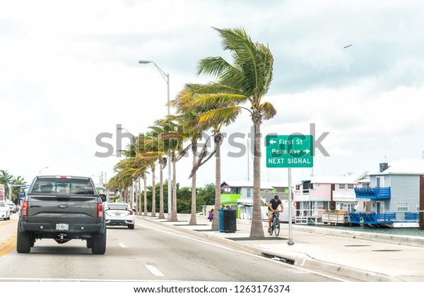 Key West, USA - May 1, 2018: Overseas highway road,\
US1, cars traffic signs for Palm avenue, first street, next signal,\
people walking, biking on bikes, palm trees row in Florida keys\
city, urban view