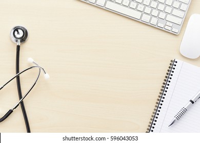 Keyboard, mouse, lined notepad, pen, and stethoscope on light colored wood desk surface.