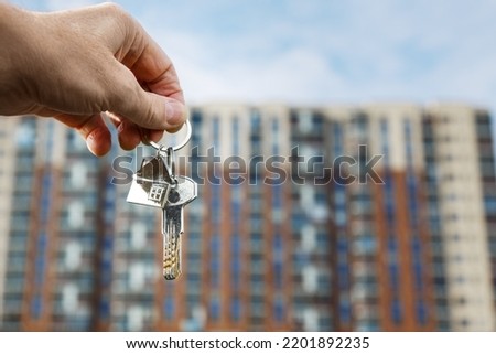 The keys to the lock with a keychain in the form of a house in the hand of a man against the background of a multi-storey residential building. Purchase, rent, insurance of a house, apartment.