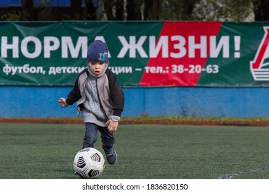 KHABAROVSK, RUSSIA - 03 Of October 2020: A Small Boy 4-6 Years Old Plays Soccer (or Football) In An Empty Stadium. Dressed For Autumn In A Hat, Jacket And Sweater