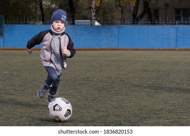 KHABAROVSK, RUSSIA - 03 Of October 2020: A Small Boy 4-6 Years Old Plays Soccer (or Football) In An Empty Stadium. Dressed For Autumn In A Hat, Jacket And Sweater