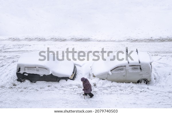 Khabarovsk, Russia - November 23, 2021: Top\
view of two cars buried under snow and an elderly woman walking on\
the snow-covered footpath after the snow\
blizzard