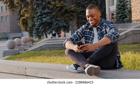 Kharkov, Ukraine - 08.09.2021: Happy African American Guy Man Sitting In City Outdoors Looking At Mobile Phone Looks In Distance Waving Hello Greeting Friend Inviting Come Here Welcome Invite