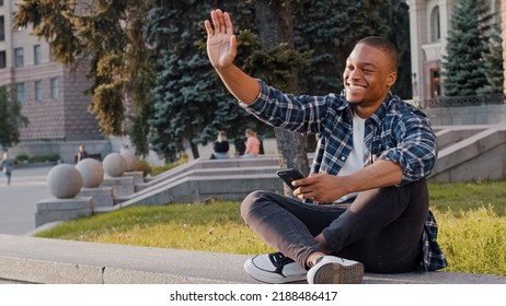 Kharkov, Ukraine - 08.09.2021: Happy African American Guy Man Sitting In City Outdoors Looking At Mobile Phone Looks In Distance Waving Hello Greeting Friend Inviting Come Here Welcome Invite