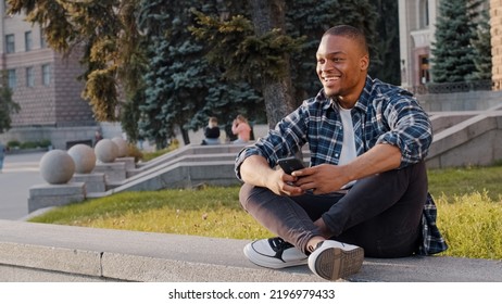 Kharkov, Ukraine - 08.09.2021: Happy African American Guy Man Sitting In City Outdoors Looking At Mobile Phone Looks In Distance Waving Hello Greeting Friend Inviting Come Here Welcome Invite Gesture