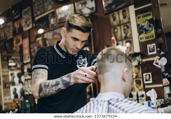 KHARKOV, UKRAINE - APRIL 17, 2019: Tattooed\
man barber in barbershop shear hair electric car to young guy for\
short hairstyle.
