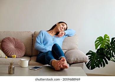 Kick Back And Relax Concept. Young Beautiful Brunette Woman With Blissful Facial Expression Alone On The Couch With Her Bare Feet On Coffee Table. Portrait Of Relaxed Female Resting At Home.