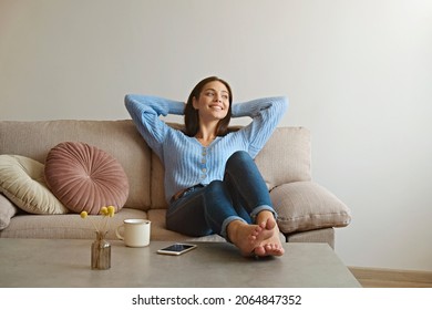 Kick back and relax concept. Young beautiful brunette woman with blissful facial expression alone on the couch with her bare feet on coffee table. Portrait of relaxed female resting at home.